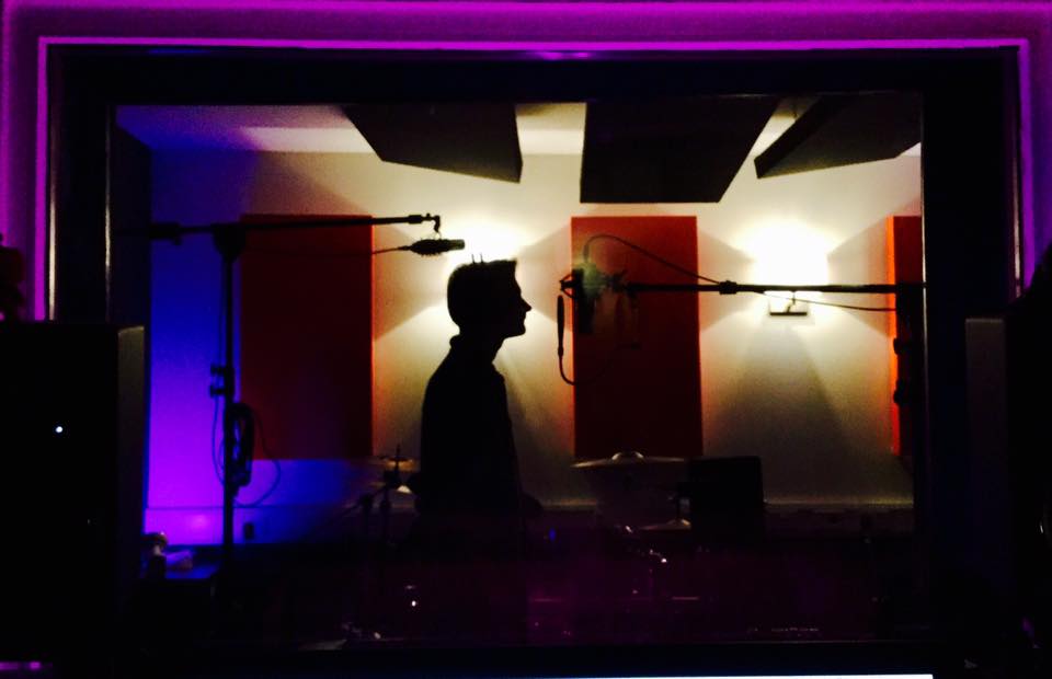 Jon Roy in silhouette at the microphone in a recording studio, lit in purple and amber